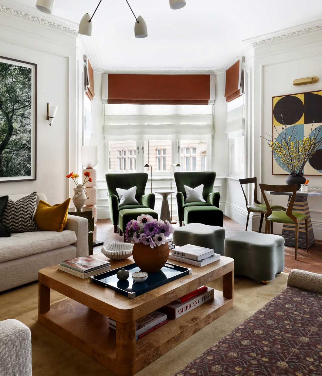 A stylish living room featuring a beige sofa, green armchairs, a wooden coffee table with books and a vase, large windows with red-orange shades, and modern art on white walls. A small dining area is in the corner.