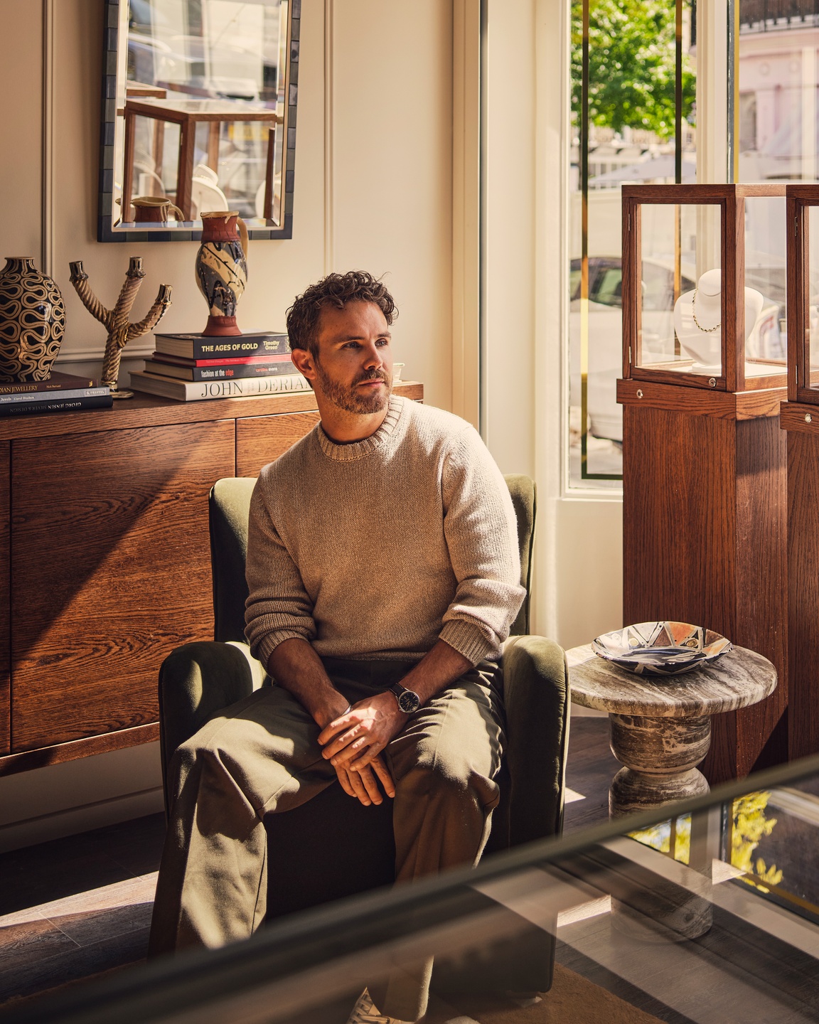 Christian Bense seated in an olive green velvet armchair in a sunlit showroom, beside oak display cabinets and a marble side table.