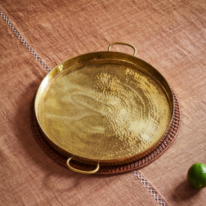 A round, hammered brass tray with two handles sits on a brown textured cloth atop a circular woven mat—a perfect accent for interior designers seeking unique finds. A green lime rests nearby