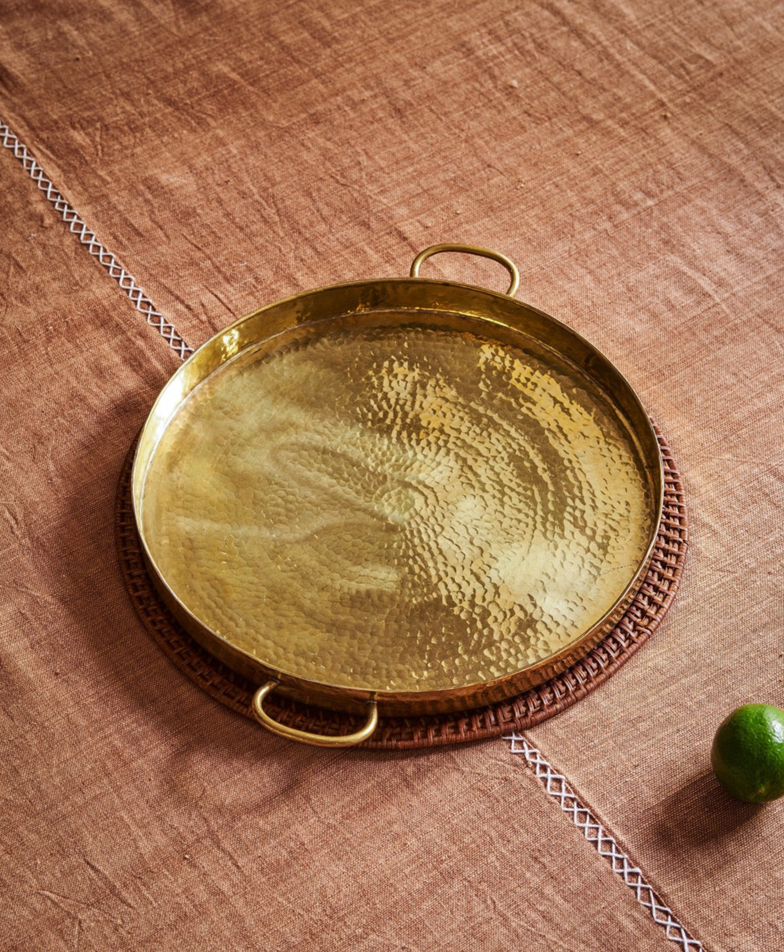 A round, hammered brass tray with two handles sits on a brown textured cloth atop a circular woven mat—a perfect accent for interior designers seeking unique finds. A green lime rests nearby