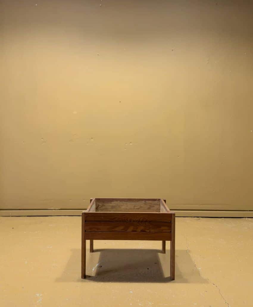 A small, empty wooden planter sits centered in a room with plain beige walls and a matching beige floor—an unadorned, minimalistic backdrop perfect for showcasing unique finds or inspiring interior designers seeking timeless home style.