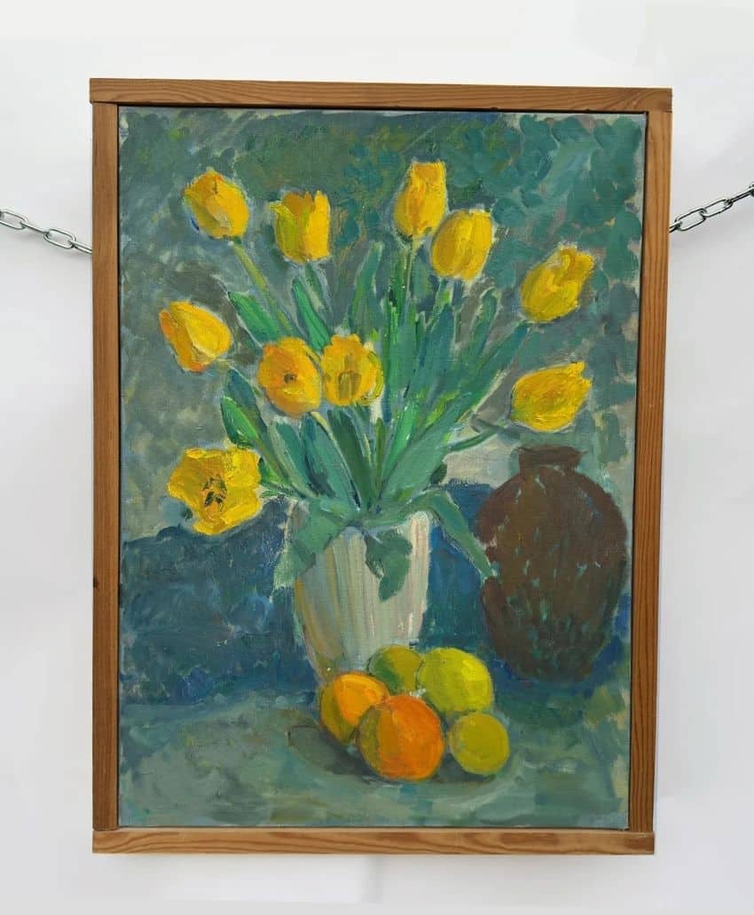 A framed painting of yellow tulips in a white vase, with oranges and green apples on a surface in front, and a dark brown jug in the background. The background is painted in blue and green tones.