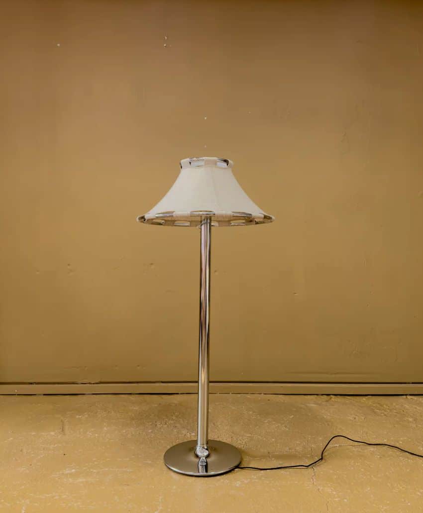 A tall floor lamp with a silver metal base and stem, topped with a cream-colored fabric lampshade, stands against a plain beige wall on a matching beige floor. A black electrical cord trails from the base.