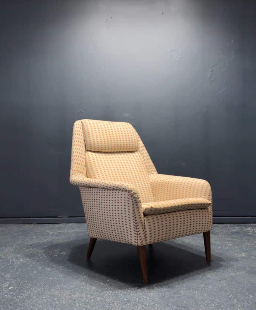 A vintage armchair with patterned upholstery and wooden legs sits on a concrete floor against a plain dark grey wall.