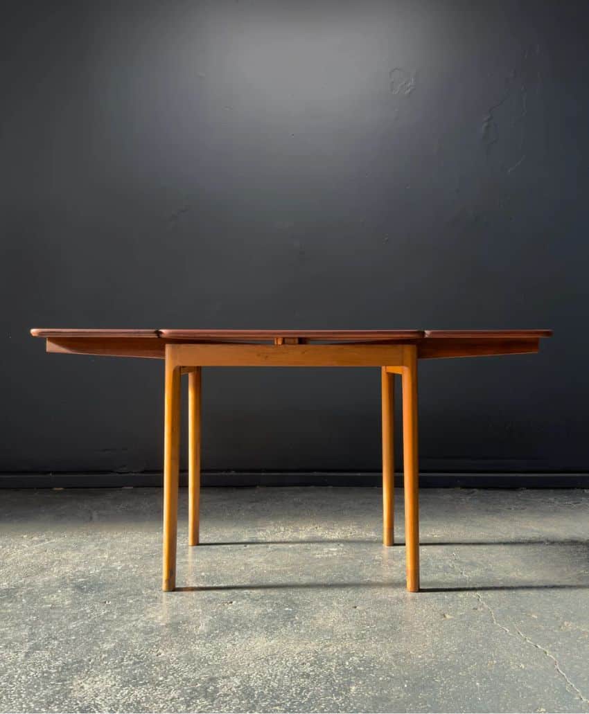 A wooden table with light-colored legs and a darker tabletop is set against a plain dark gray wall and concrete floor. The table has a simple, mid-century modern design.