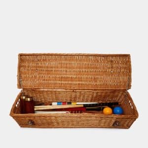 A rectangular wicker basket with an open lid contains croquet mallets, coloured balls, and other croquet equipment neatly arranged inside.