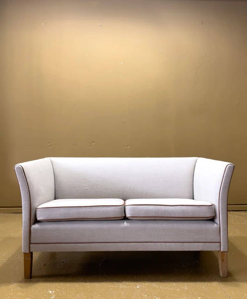 A small, light-colored loveseat with wooden legs sits against a plain tan wall and brown floor in a minimalist setting.