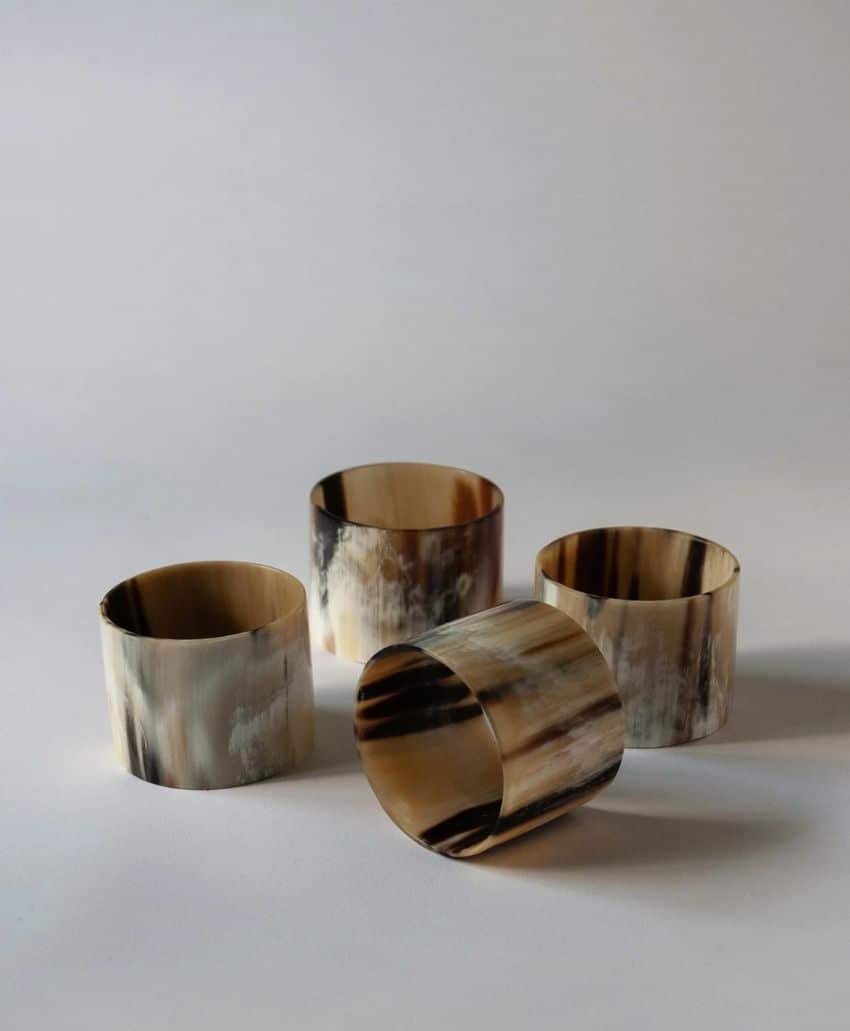 Four cylindrical napkin rings with a marbled brown, black, and cream pattern are arranged on a neutral surface. One ring is lying on its side while the others are standing upright.