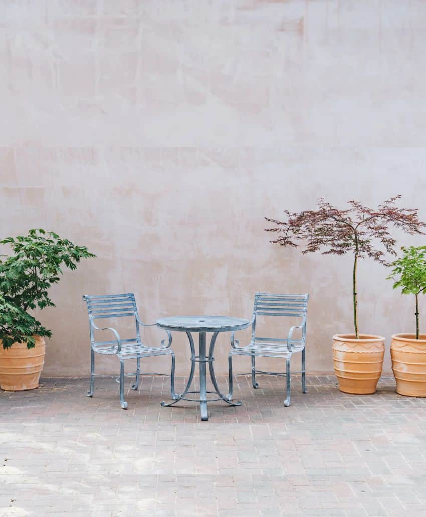 Two empty metal chairs and a round table sit on a brick patio, flanked by three potted plants against a light, textured wall.