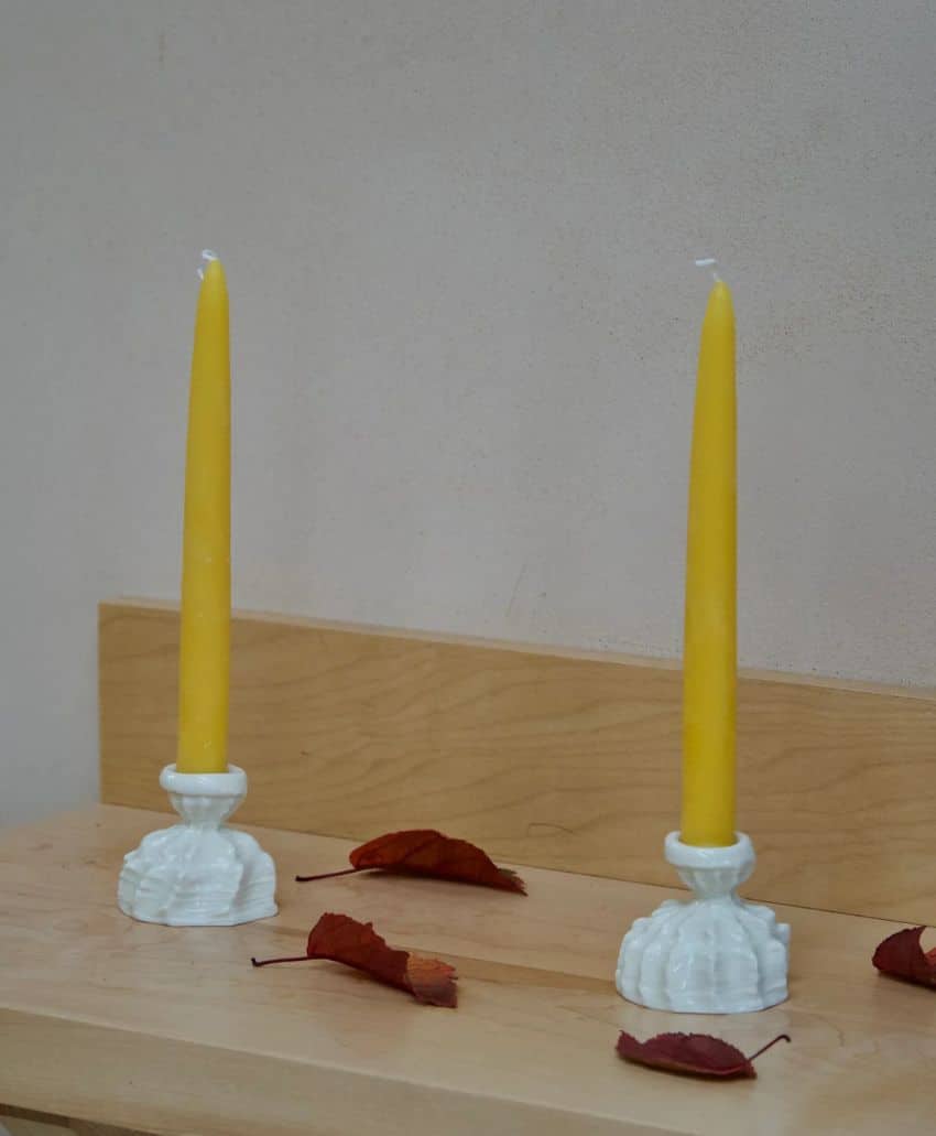 Two yellow taper candles in white holders are placed on a light wooden surface. Several dried brown leaves are scattered around the candles. The background is plain and light-colored.