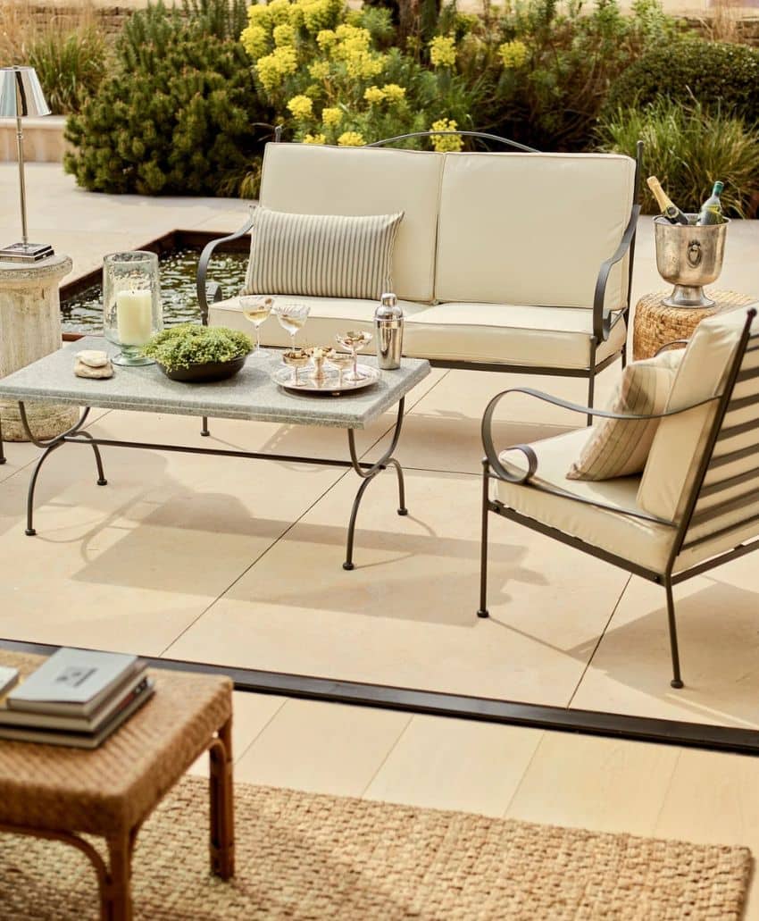 Outdoor patio seating area with a cream-colored sofa and chair, glass-top coffee table set with drinks and snacks, and a wicker tray on a side table, surrounded by lush plants and a small pond.