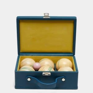 A blue leather box with a yellow interior, open to reveal six light wooden balls with green lines and one small pink ball inside.