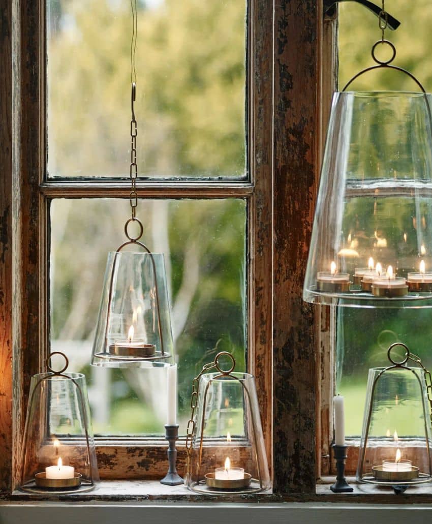 Glass lanterns and candle holders with lit candles are arranged on a rustic wooden windowsill and hung in front of a window, with greenery visible outside.