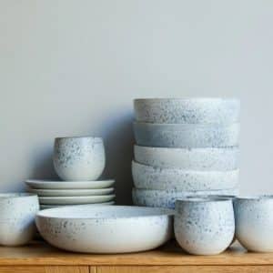 A set of white, speckled ceramic bowls and plates are stacked neatly on a light wooden cabinet against a plain, light grey wall.