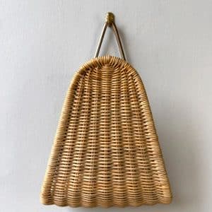 A woven wicker basket with a curved top and scalloped bottom edge hangs from a metal hook on a plain white wall.