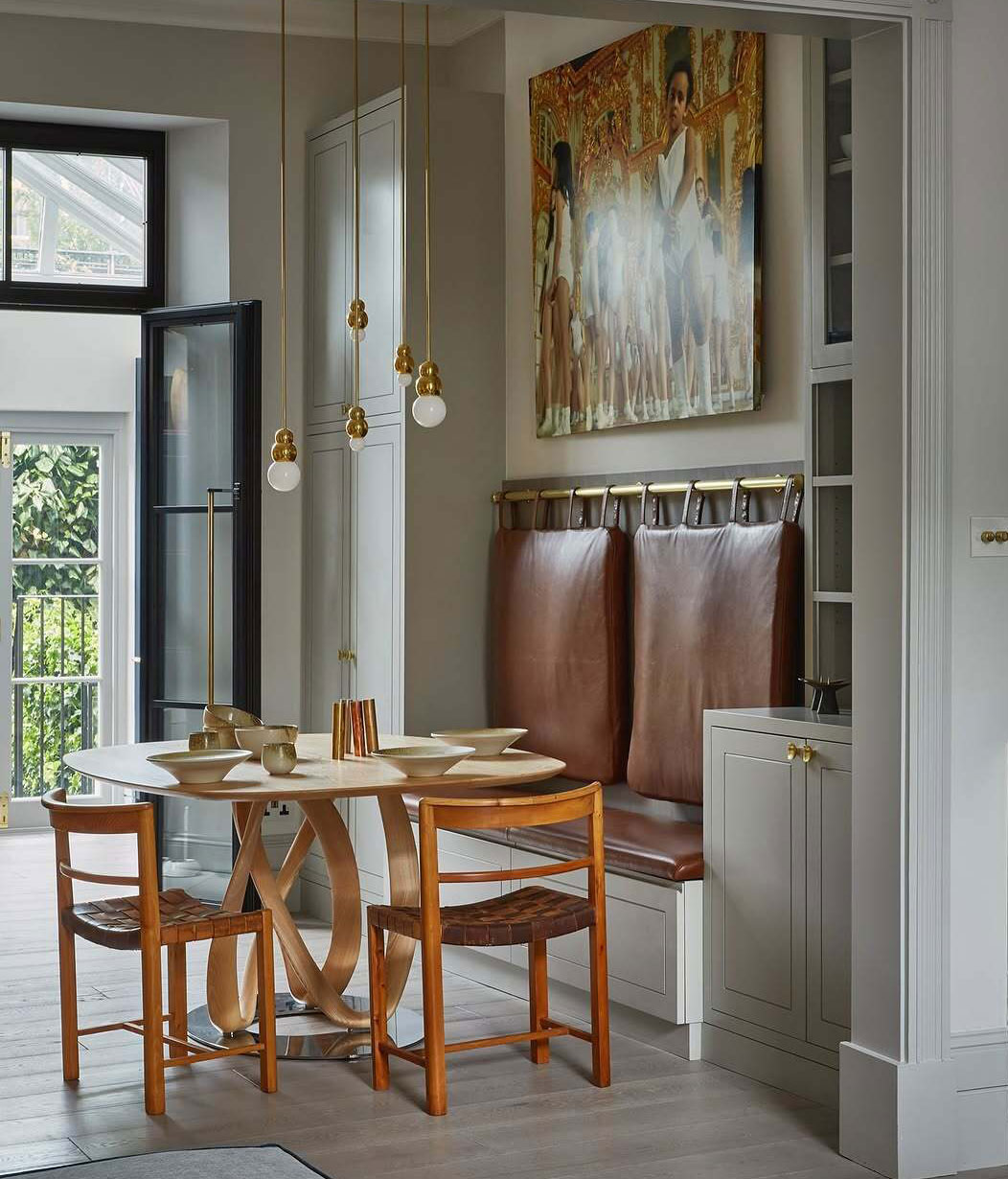 A cozy dining nook with a modern round table, four wooden chairs, leather bench seating, pendant lights, and a large abstract painting above the bench. Light streams in from a windowed door, brightening the stylish space.