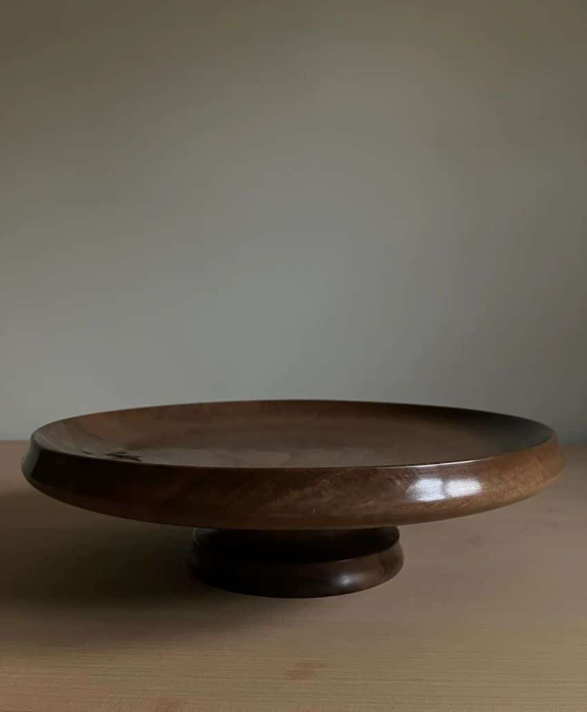 A wooden pedestal cake stand with a smooth, round surface and short base, placed on a light wooden table against a plain, neutral background.