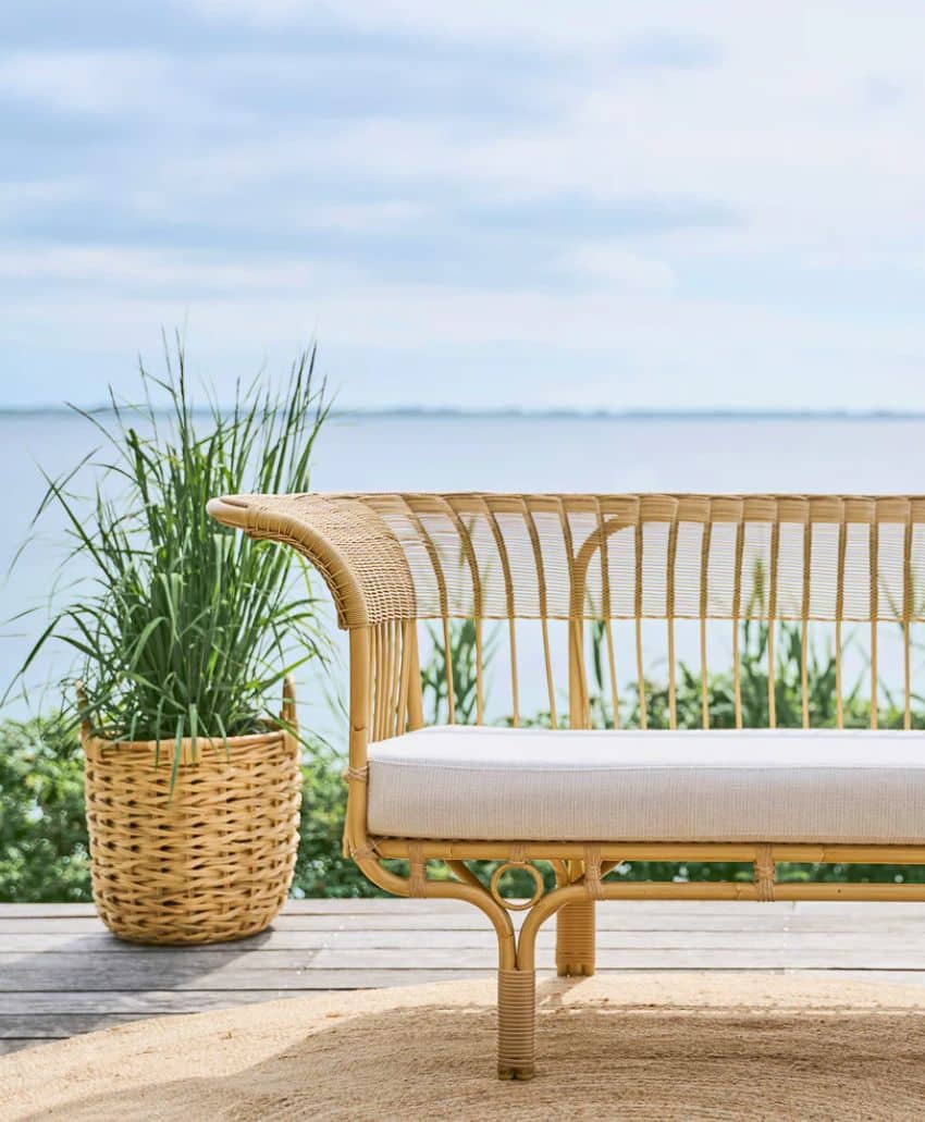 A wicker sofa with a white cushion sits on a wooden deck overlooking water, with a potted green plant beside it. The background features a calm, cloudy sky and lush greenery.