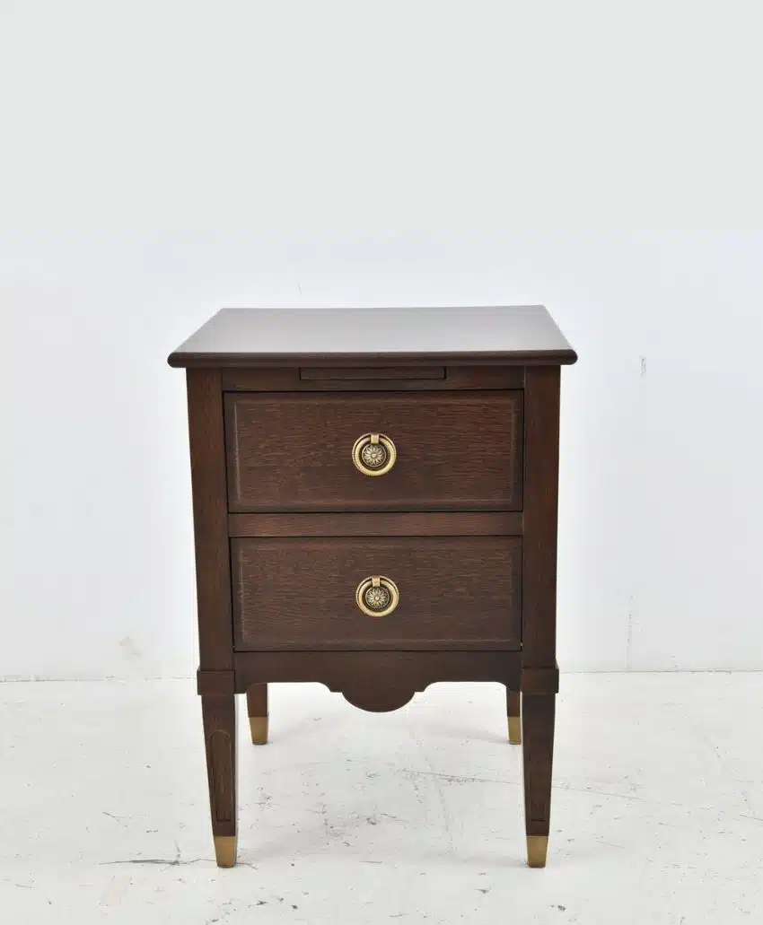 A dark wooden nightstand with two drawers, each featuring a round brass handle. The nightstand has tapered legs with brass caps and a simple, classic design, standing against a plain white background.