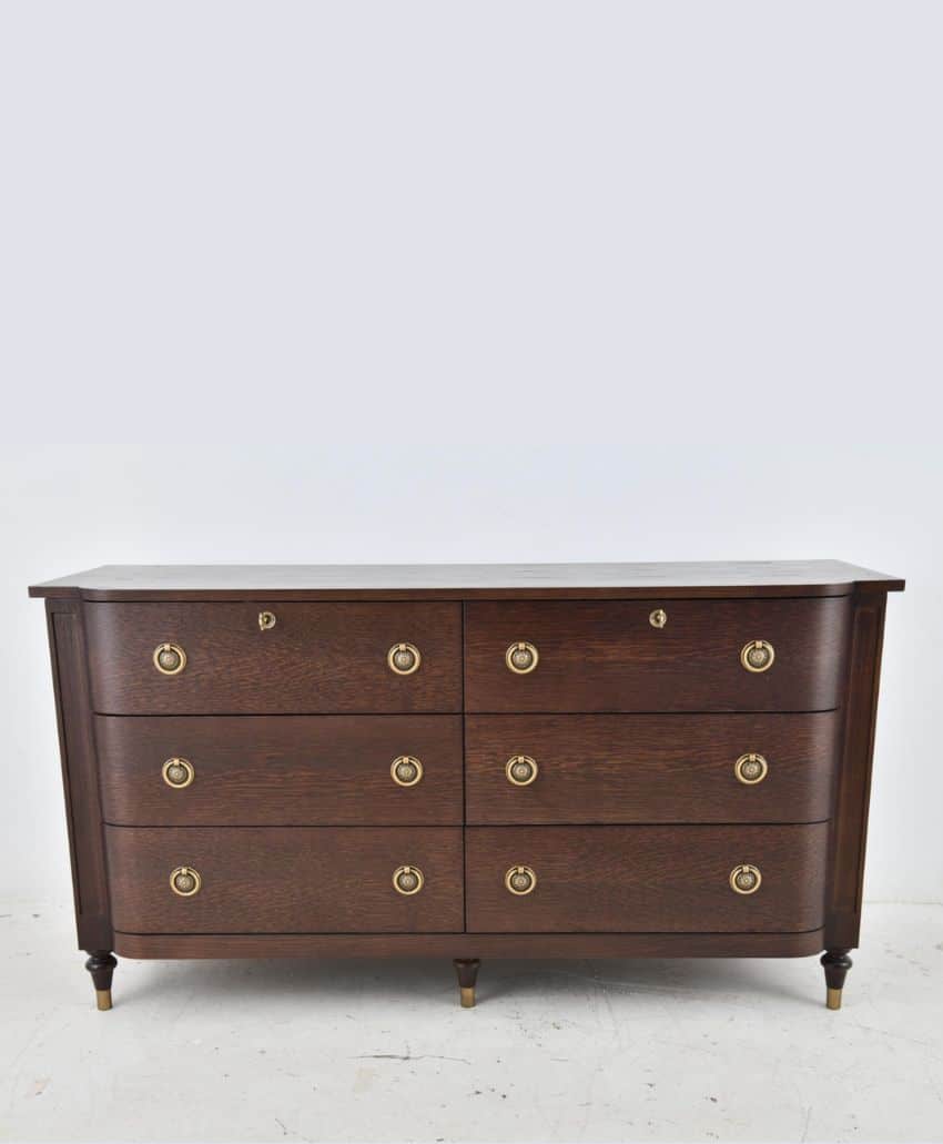 A dark wood dresser with six drawers, each featuring two round brass handles, stands against a plain light gray background. The dresser has tapered legs and a smooth, polished surface.