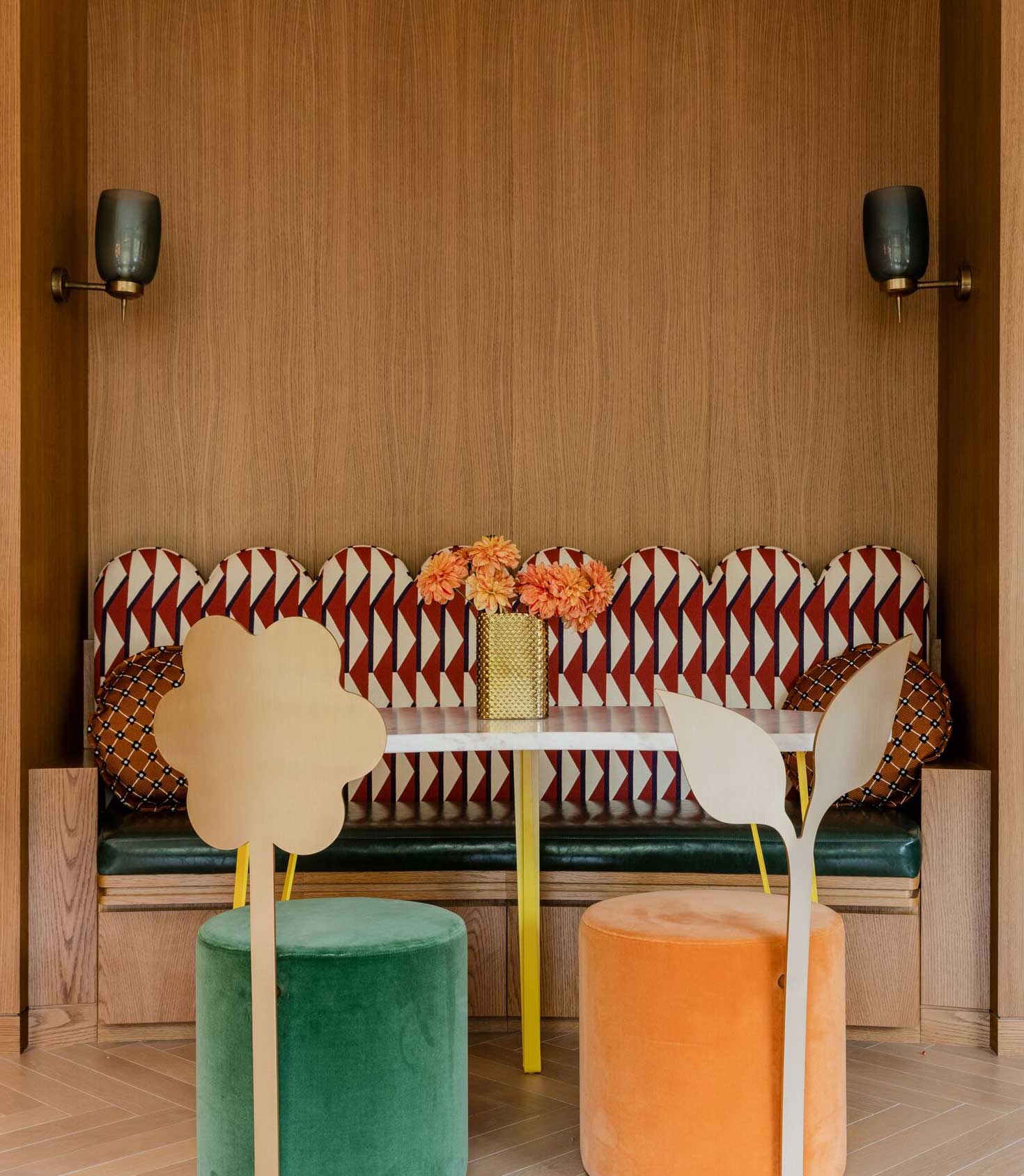 A modern dining room with a light wood table, tan chairs, a floral arrangement, a yellow and black sideboard, and a wall mural featuring abstract green trees and plants. A contemporary pendant light hangs above the table.