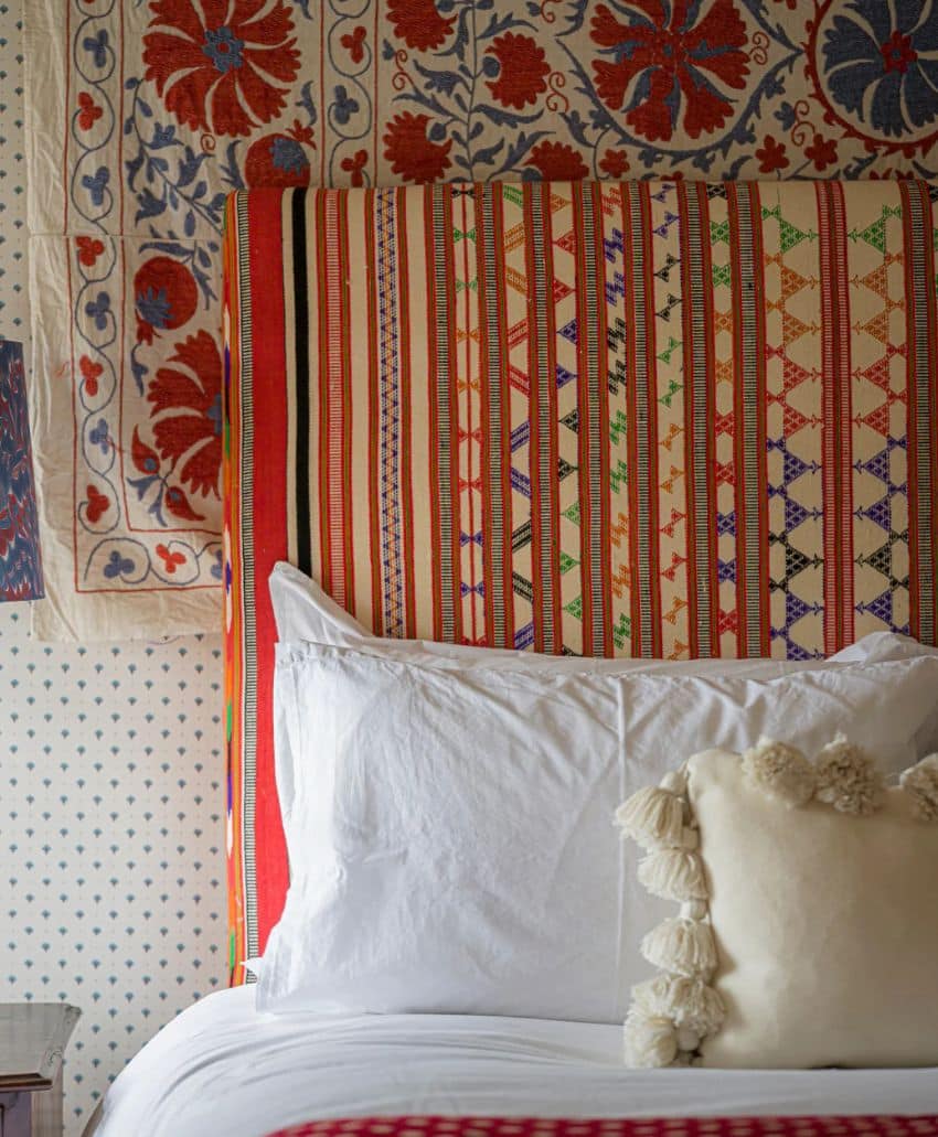 A bed with white pillows, one featuring pom-pom tassels, sits against a headboard upholstered in colorful, striped and geometric fabric. Tapestries with red floral and blue designs hang on the wall behind.