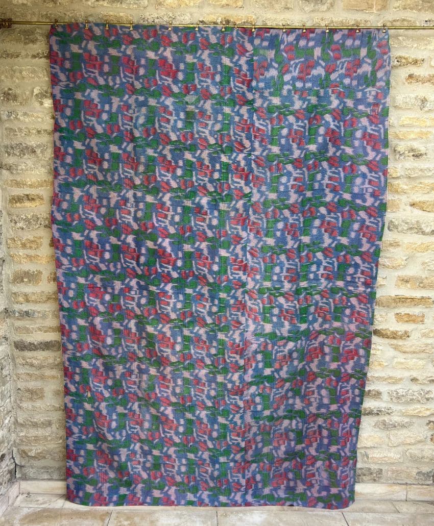 A large, rectangular fabric with an abstract, multicolored pattern in blue, green, red, and purple tones is hanging against a rough stone wall background.
