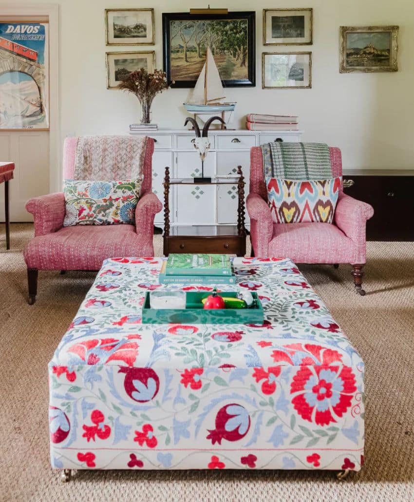 Two pink armchairs with patterned cushions sit behind a floral ottoman table in a cozy living room. A white sideboard, books, framed art, and a model sailboat decorate the background.