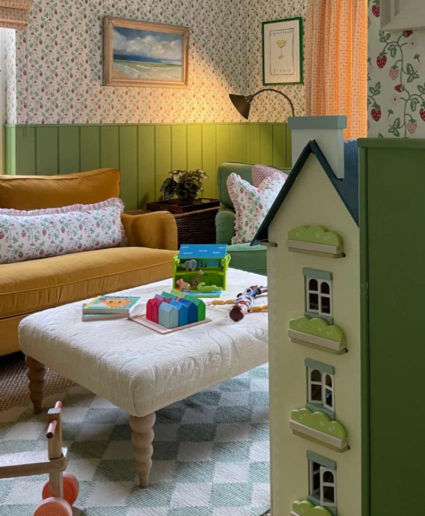 A cozy, vintage-style living room with floral wallpaper, a yellow sofa, patterned cushions, a white ottoman with wooden toys, and a green-and-white dollhouse in the foreground. Soft, warm lighting completes the scene.