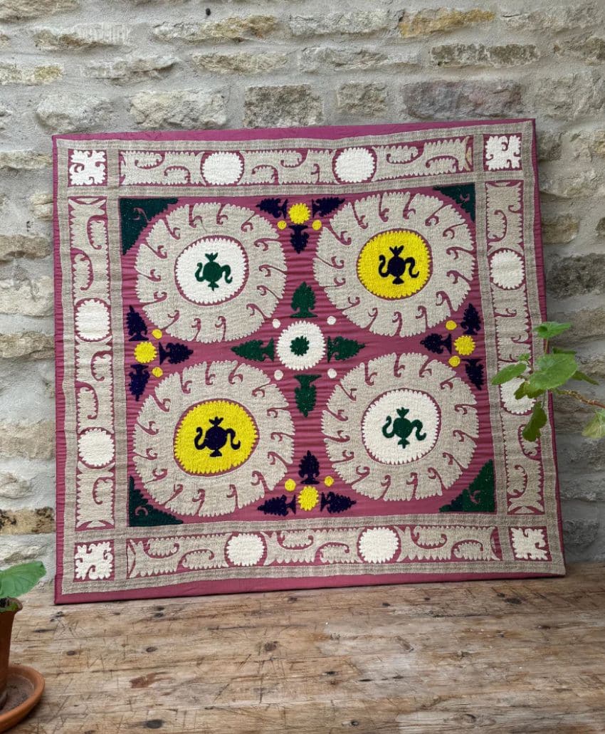 A colorful, handmade cloth board for the game Parchisi with intricate designs, circular patterns, and four central yellow and green sections, set against a stone wall with some greenery visible.