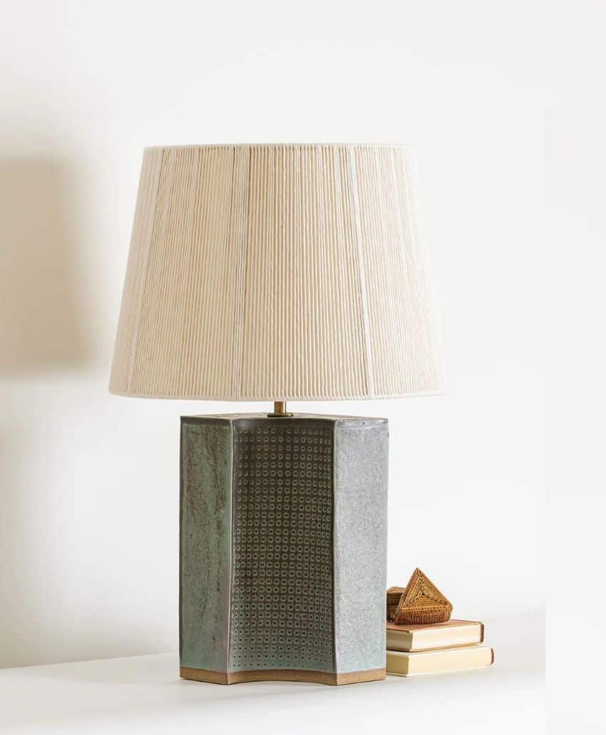 A modern table lamp with a textured grey geometric base and a pleated white lampshade sits on a white surface next to a small stack of books and a decorative item.