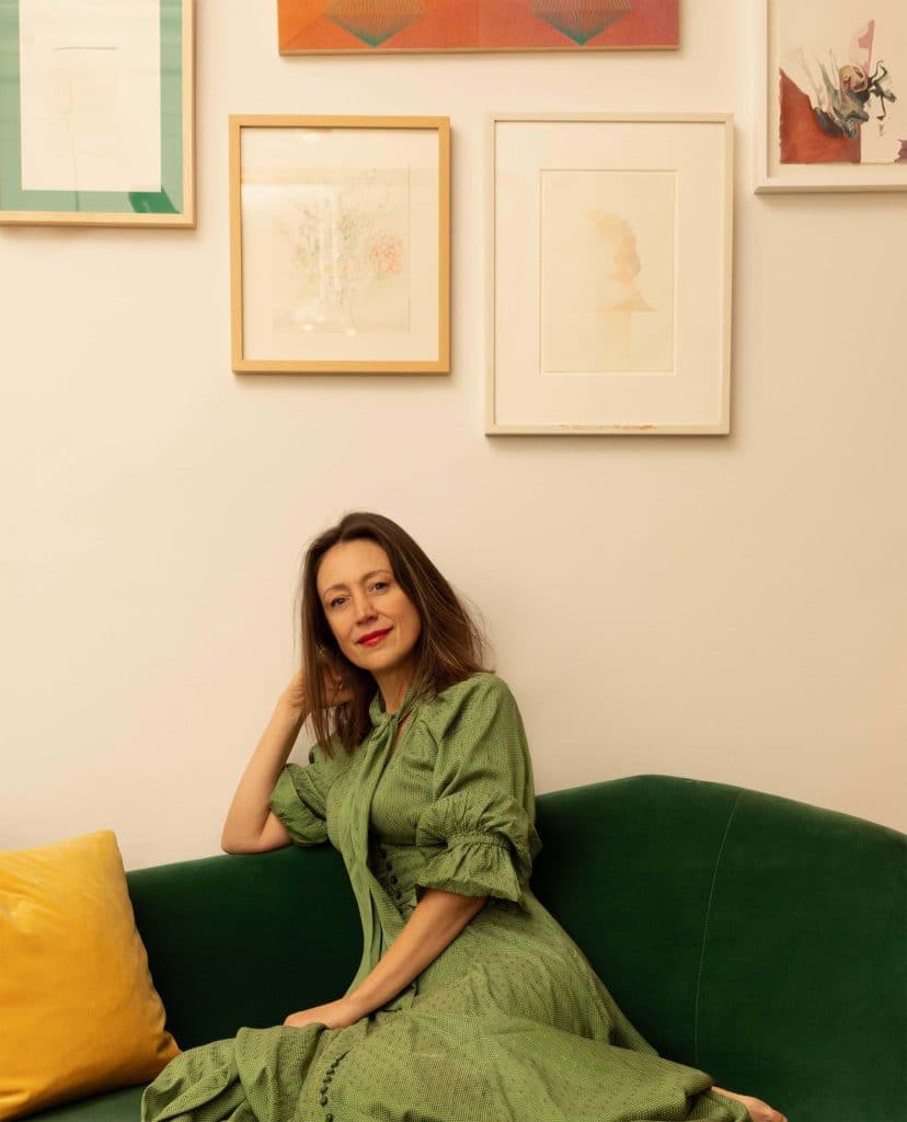 A woman in a green dress sits on a dark green sofa with a yellow cushion, smiling, against a white wall decorated with framed artwork and photos.