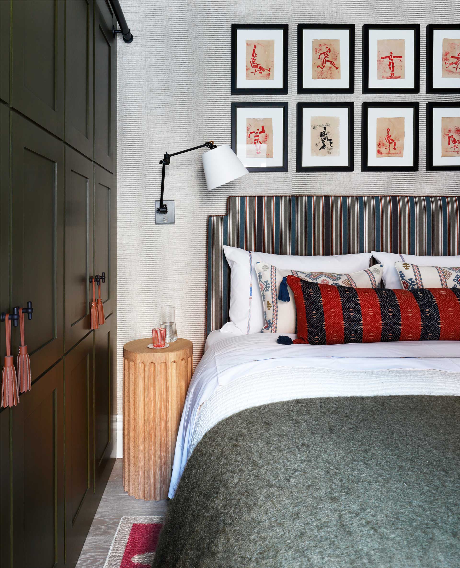 A cozy bedroom with a striped headboard, colorful pillows, and a green blanket on the bed. A small wooden side table holds a cup and glass. Nine framed art pieces hang above the bed next to olive green built-in wardrobes.