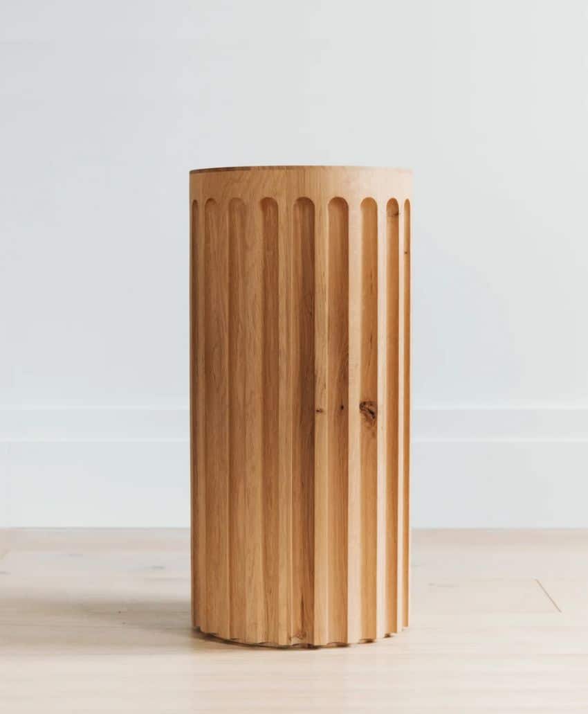 A cylindrical wooden stool with vertical grooves stands on a light wooden floor against a plain white wall. The stool has a smooth, natural finish and a minimalist design.