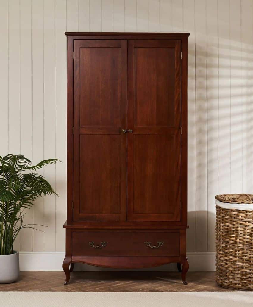 Tall, dark wooden armoire with two paneled doors and a lower drawer, standing on curved legs. It is placed against a cream-coloured wall near a green plant and a woven laundry basket.