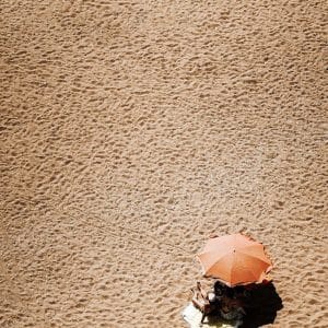 Two people sit under an orange umbrella on a sandy beach, surrounded by footprints and textured sand, with much empty space around them.