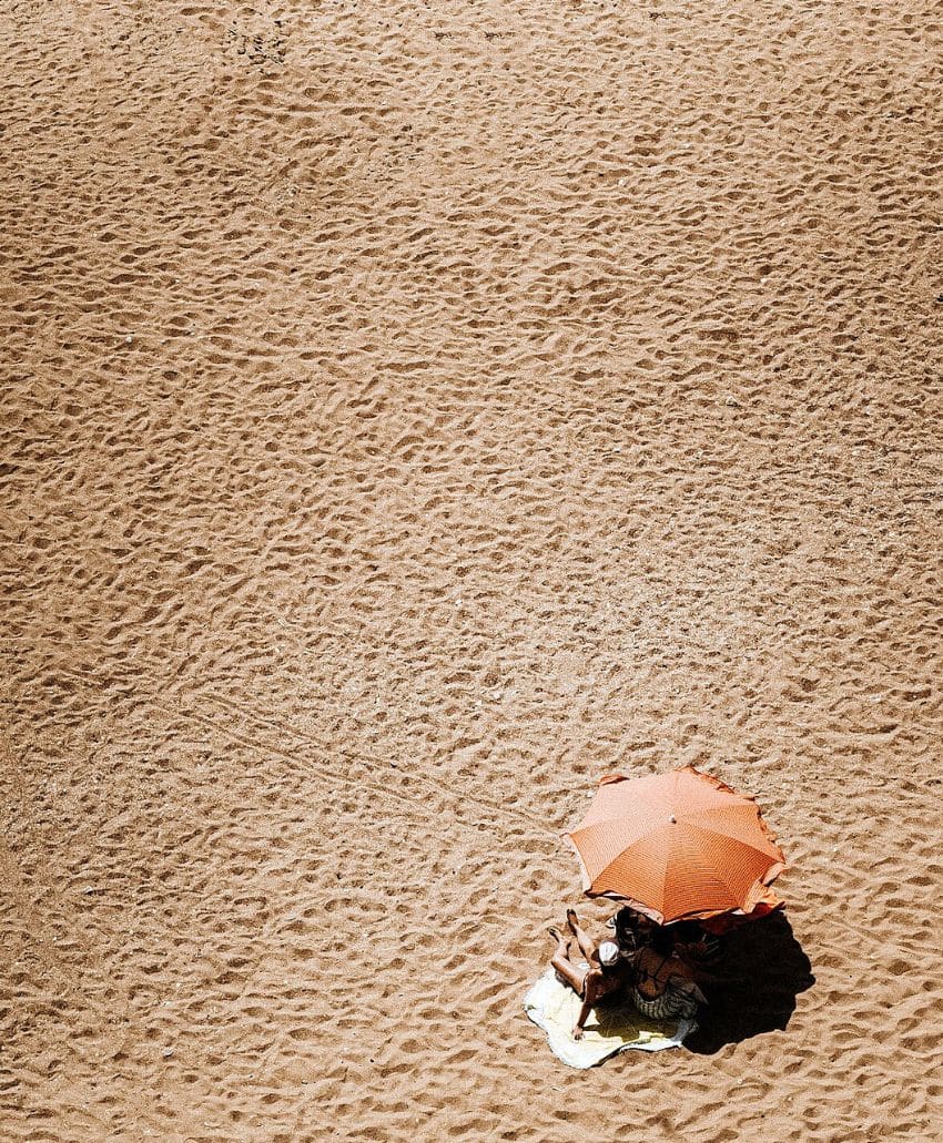 Two people sit under an orange umbrella on a sandy beach, surrounded by footprints and textured sand, with much empty space around them.