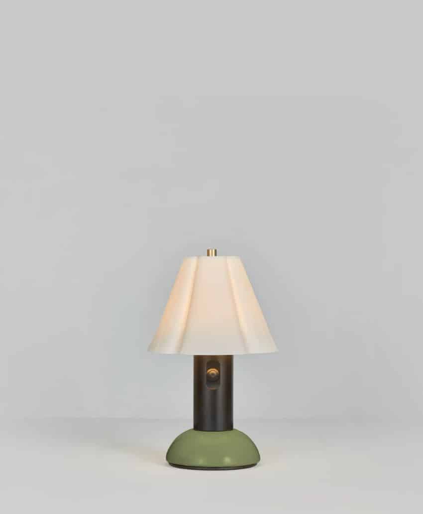 A small table lamp with a white shade, a dark cylindrical base, and a round green bottom, set against a plain light gray background.