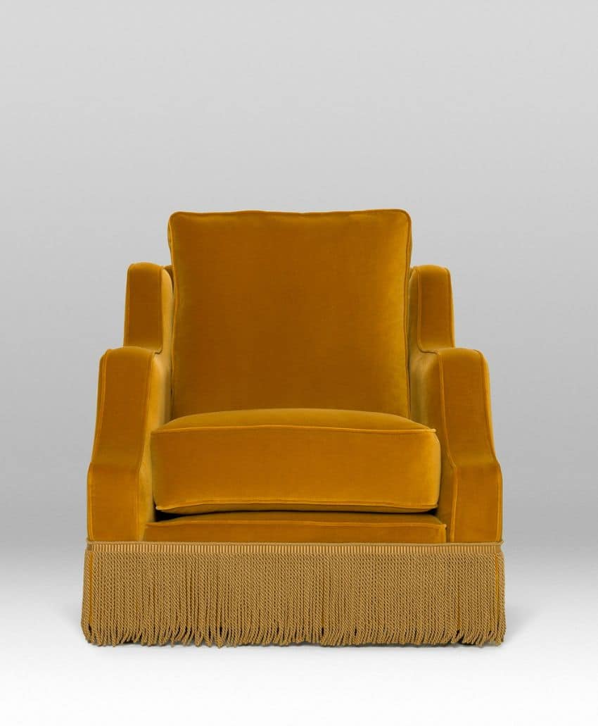 A mustard yellow velvet armchair with a boxy shape and wide armrests, featuring a decorative fringe at the base, set against a plain light gray background.