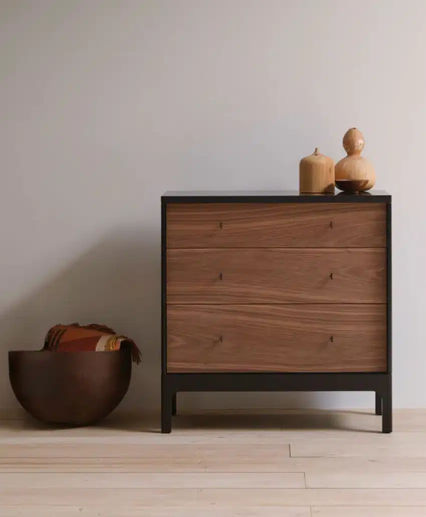 A modern wooden dresser with three drawers sits against a plain wall. On top are two decorative vases. To the left, a brown bowl holds neatly folded blankets. The floor is light wood.