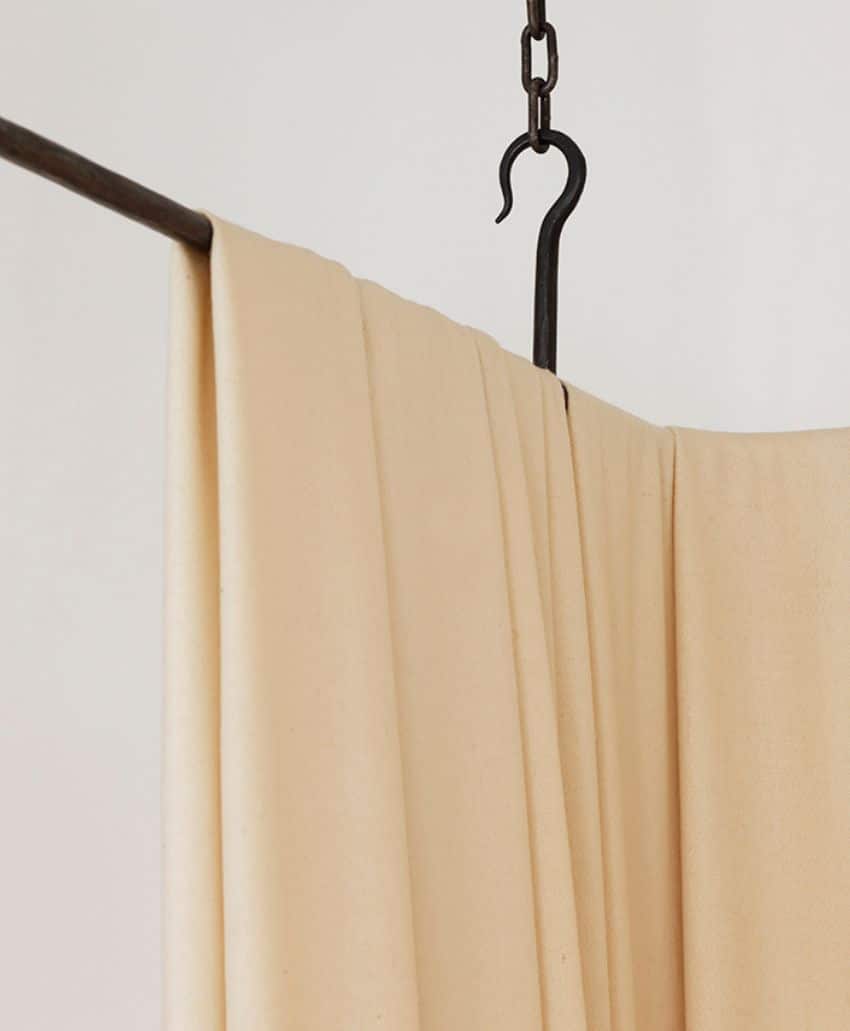 A beige curtain hangs from a dark metal rod and a large hook against a light-colored wall. The fabric drapes in soft folds, creating a minimalist and calm appearance.
