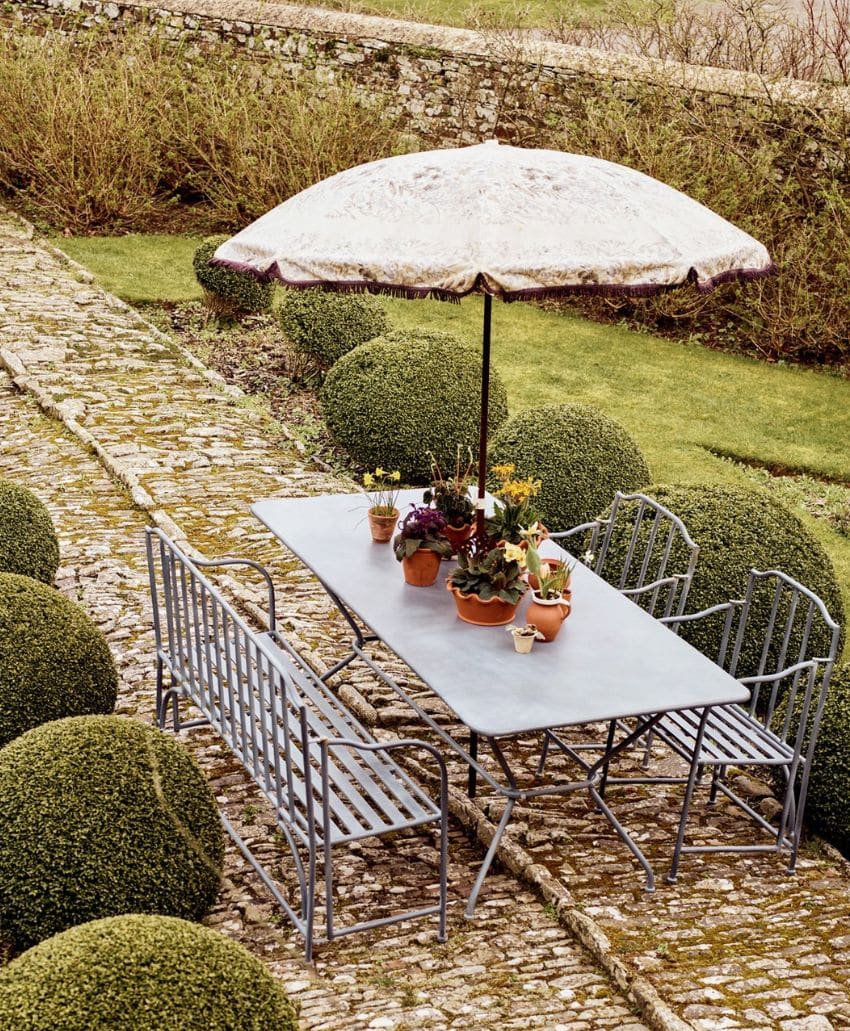 A metal outdoor table with potted plants on top, surrounded by benches and chairs, sits on a stone patio. An open parasol provides shade, and manicured bushes line the garden path nearby.
