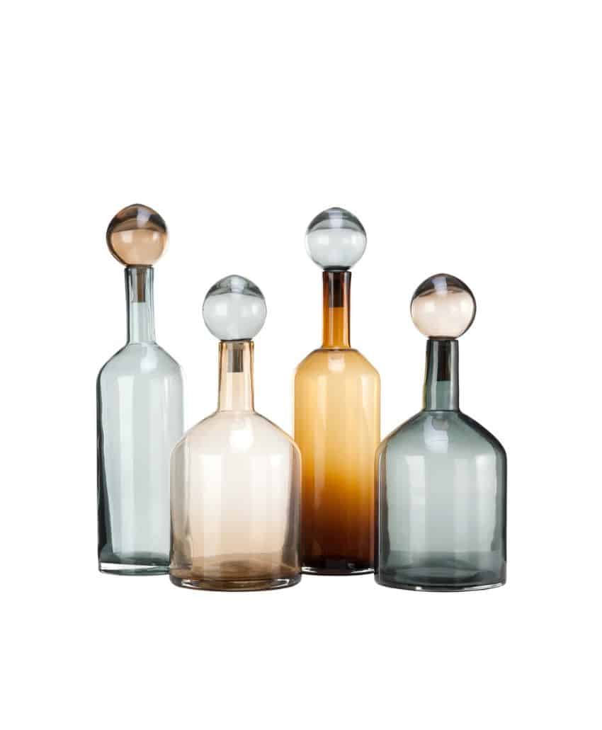 Four glass decanters with round stoppers in pastel shades of clear, amber, grey, and smoky brown, arranged in a group on a white background.
