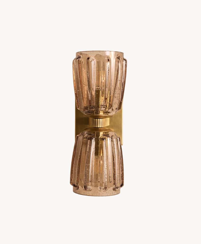 A modern wall sconce with two ribbed, amber glass shades—one facing up and one facing down—mounted on a rectangular brass base, set against a plain light background.