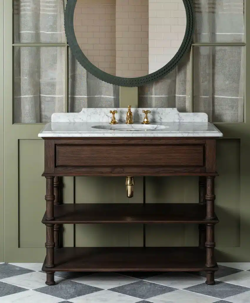 A dark wood bathroom vanity with open shelves, a white marble countertop, and gold faucet fixtures sits below a round mirror. The background features green paneling and sheer curtains, with a checkered tile floor.