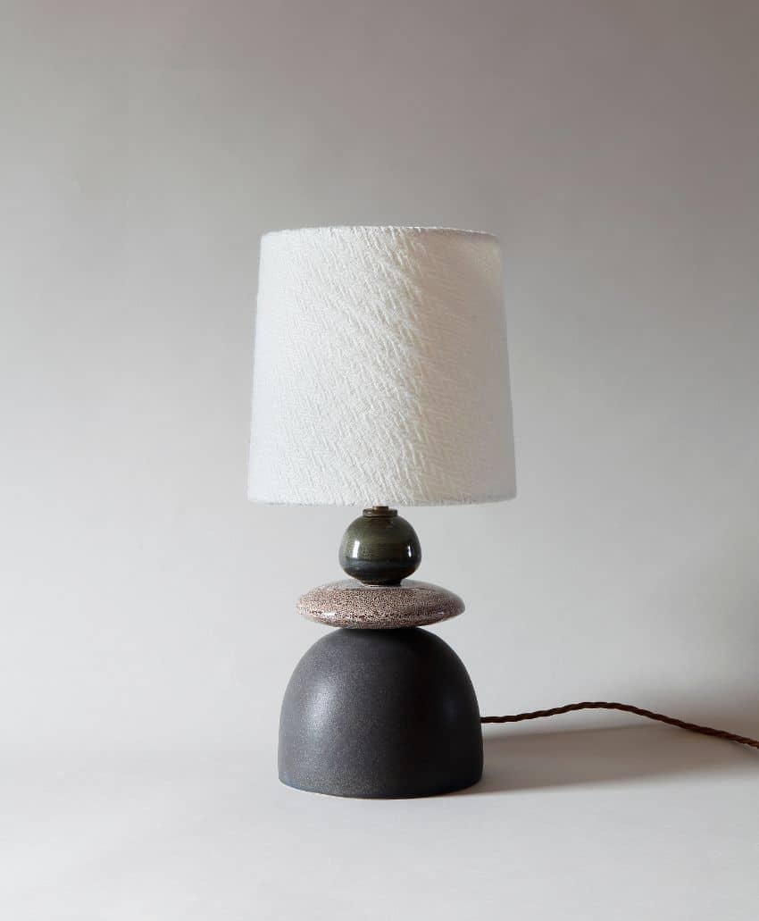 A modern table lamp with a textured white lampshade, dark round ceramic base, and decorative stone accent, displayed against a plain light background with a brown twisted power cord.