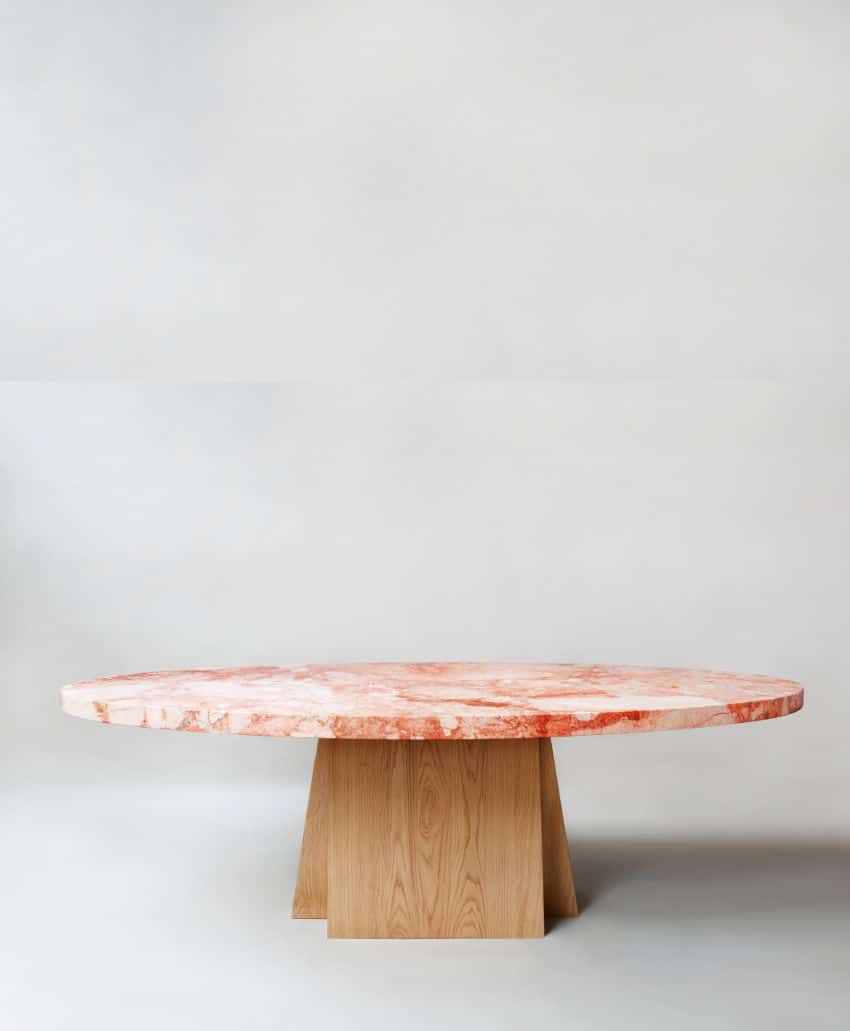 A round table with a pinkish marble top and a geometric wooden base, set against a plain, light background.