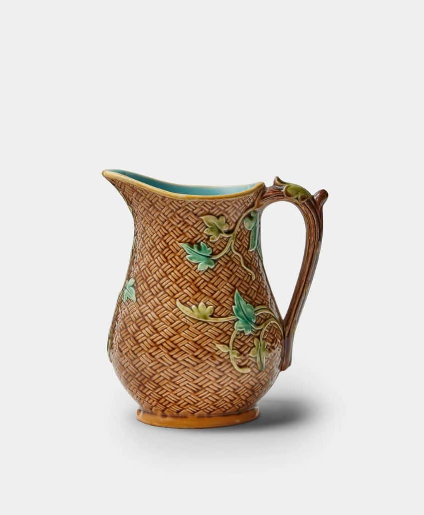 A brown ceramic pitcher with a textured, basket-weave pattern and raised green leaf and vine decorations, featuring a curved handle and a teal interior.