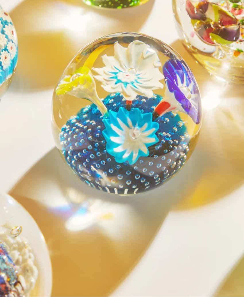 A glass paperweight with blue and white flower designs and tiny bubbles inside, resting on a white surface with soft, colorful reflections.
