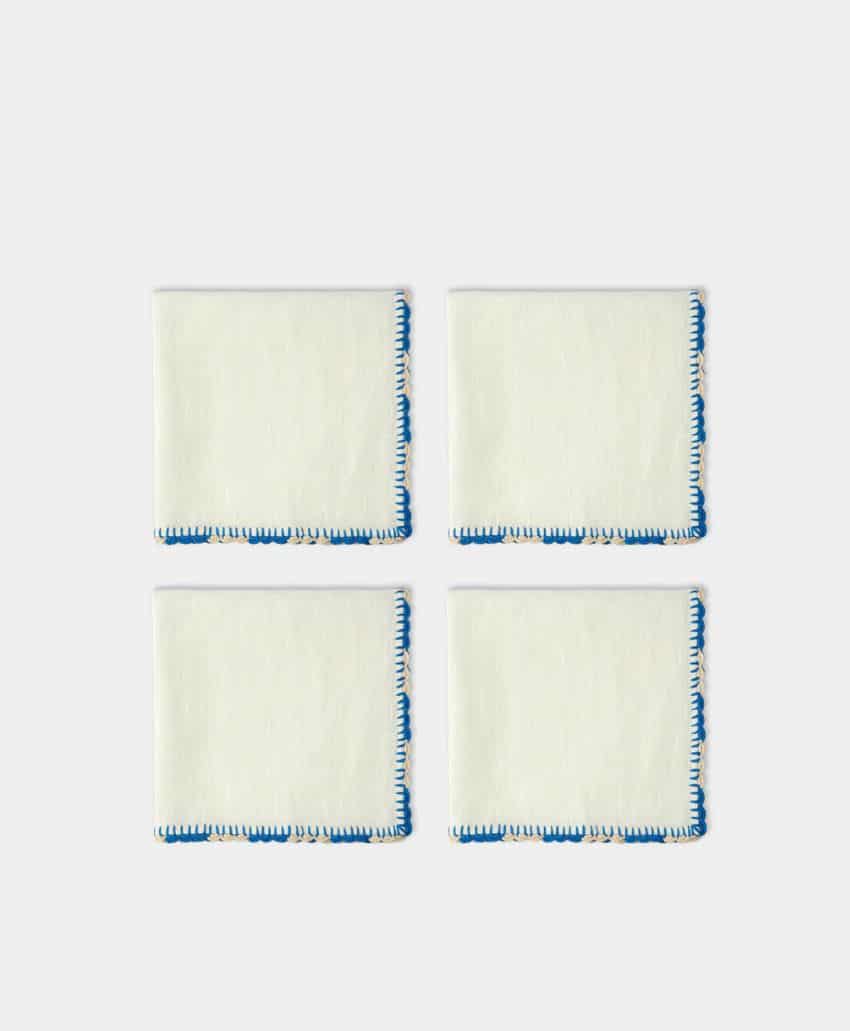 Four square white cloth napkins with blue decorative stitching along the edges, arranged in a 2x2 grid on a plain light background.